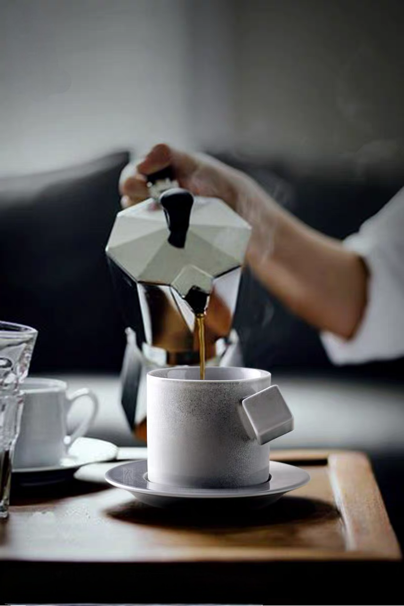 Gray ceramic cup and saucer set with espresso being poured from moka pot highlighting modern minimalist design