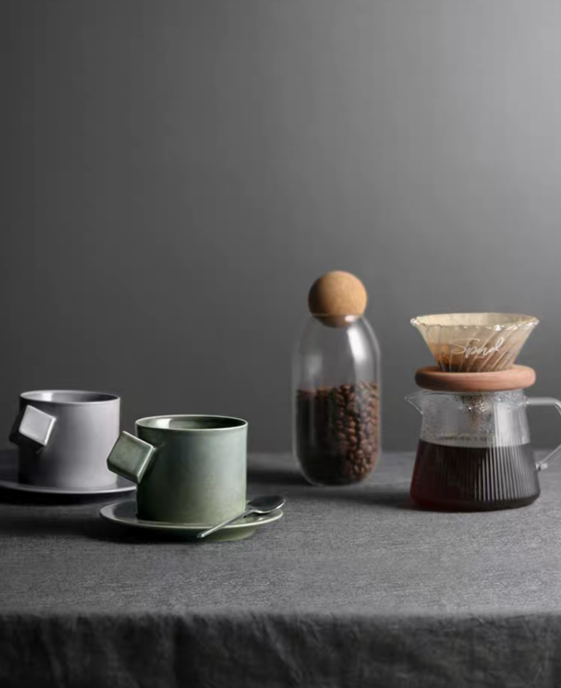 Gray and green ceramic cup and saucer set placed beside coffee brewing setup with beans and pour over dripper in lifestyle scene