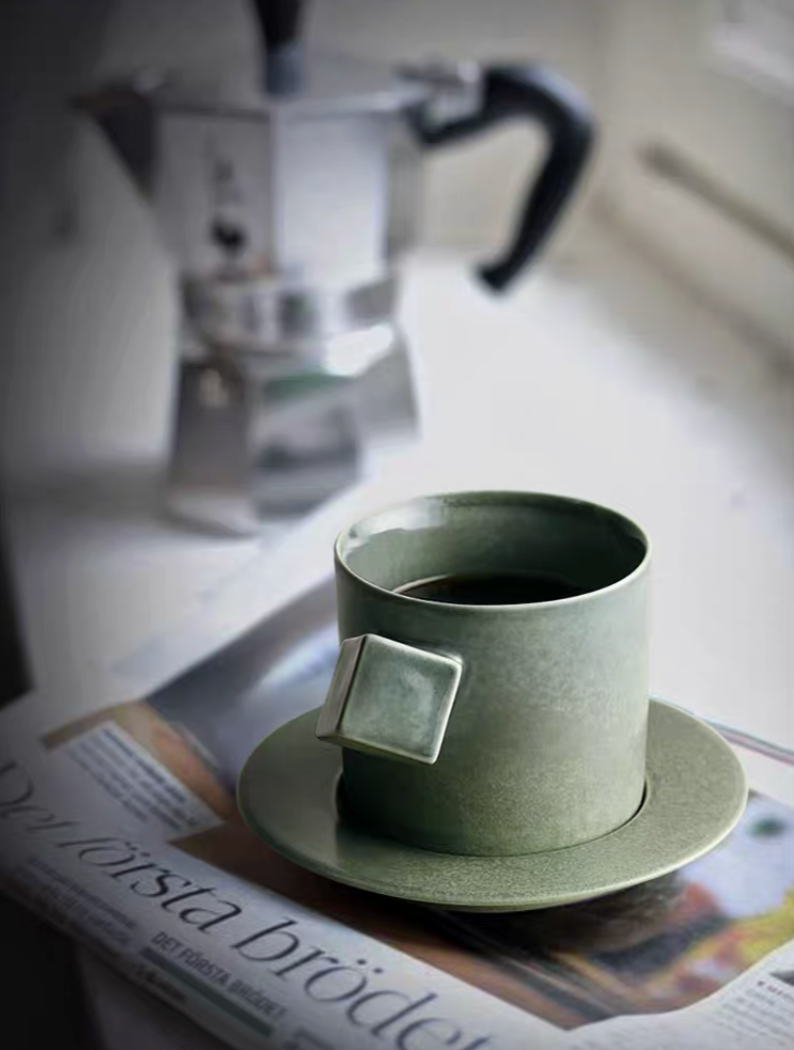 Green ceramic cup and saucer set placed on newspaper by window with coffee inside creating calm lifestyle setting