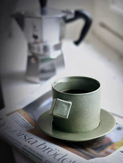 Green ceramic cup and saucer set placed on newspaper by window with coffee inside creating calm lifestyle setting