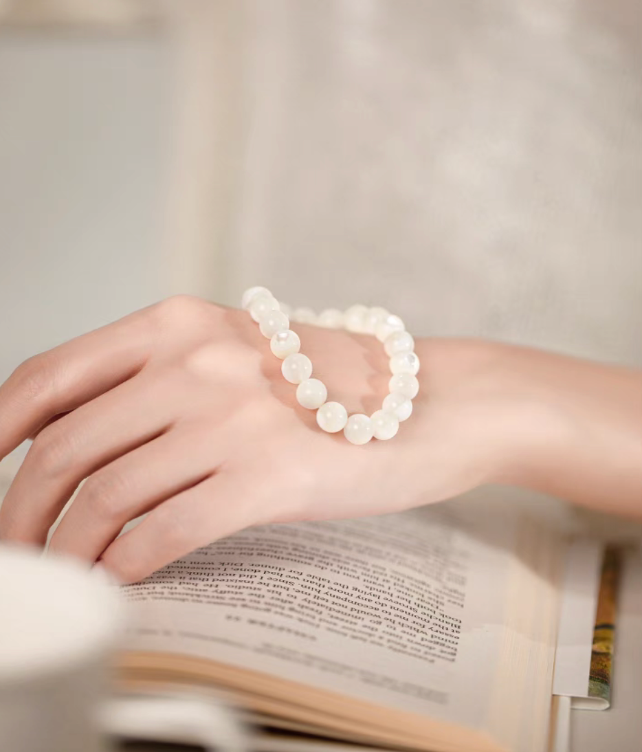 Natural white mother-of-pearl bracelet looped over a hand resting above an open book in a soft neutral setting