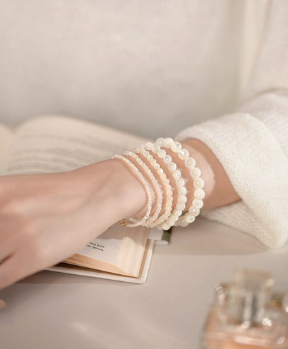 Several natural white mother-of-pearl bracelets stacked on a wrist above an open book in a soft lifestyle scene