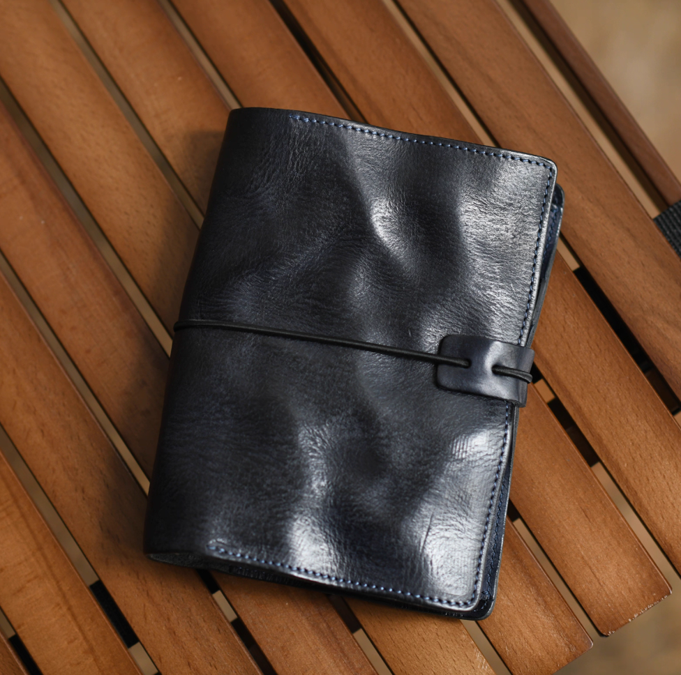 Navy blue leather refillable journal with elastic wrap placed on wooden slatted table.