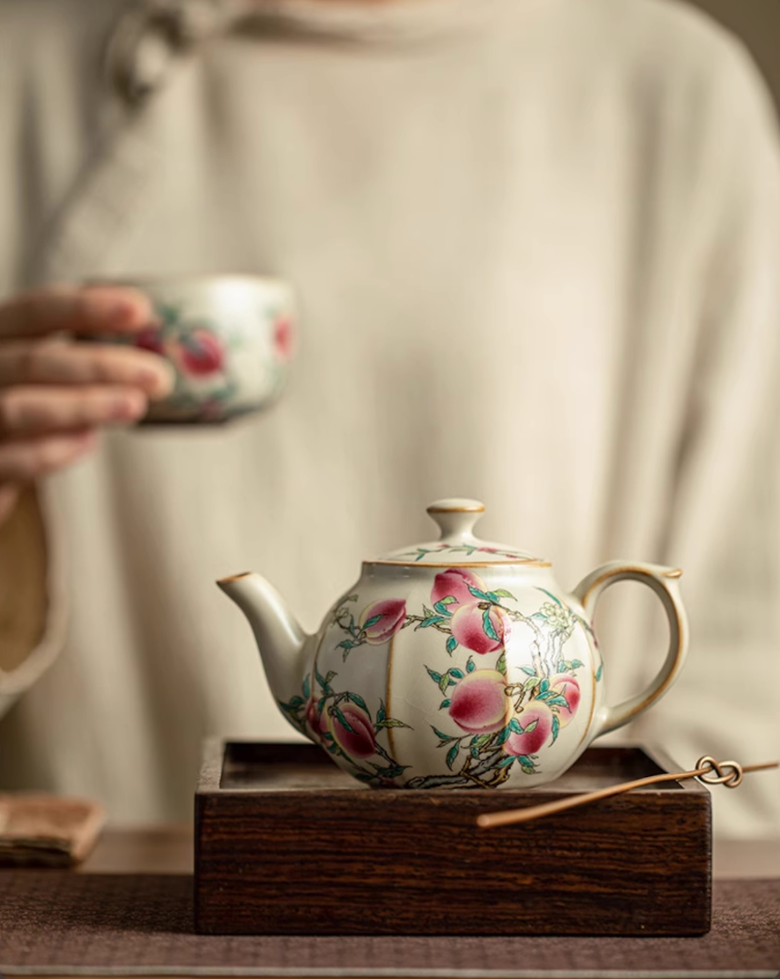 Nine Peach Ru Kiln Crackle Glazed Teapot in Nine Peach color with peach blossom pattern, front view placed on a wooden base with matching teacup in background