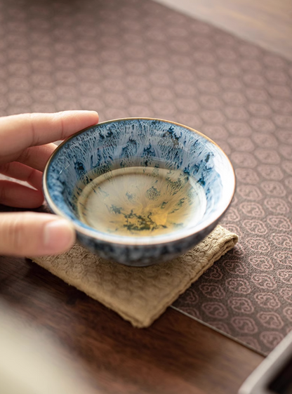 Blue kiln-glazed ceramic tea cup filled with tea placed on cloth mat showing natural glaze variation