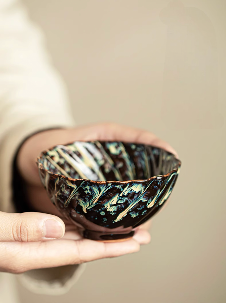 Hand holding textured dark kiln-glazed tea cup with sculpted exterior pattern and glossy ceramic finish