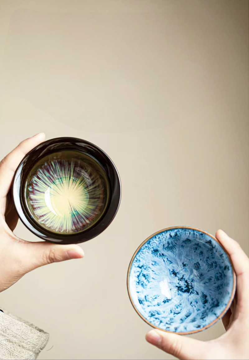 Two kiln-glazed tea cups held in hands showing contrasting interior glaze patterns in blue and multicolor finish