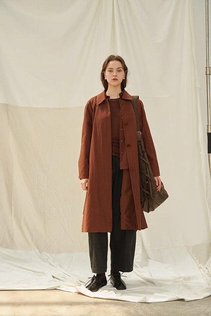Model wearing terracotta brown vintage A-line raglan trench coat full-length front view, styled with dark cropped pants and lace-up shoes against draped fabric background