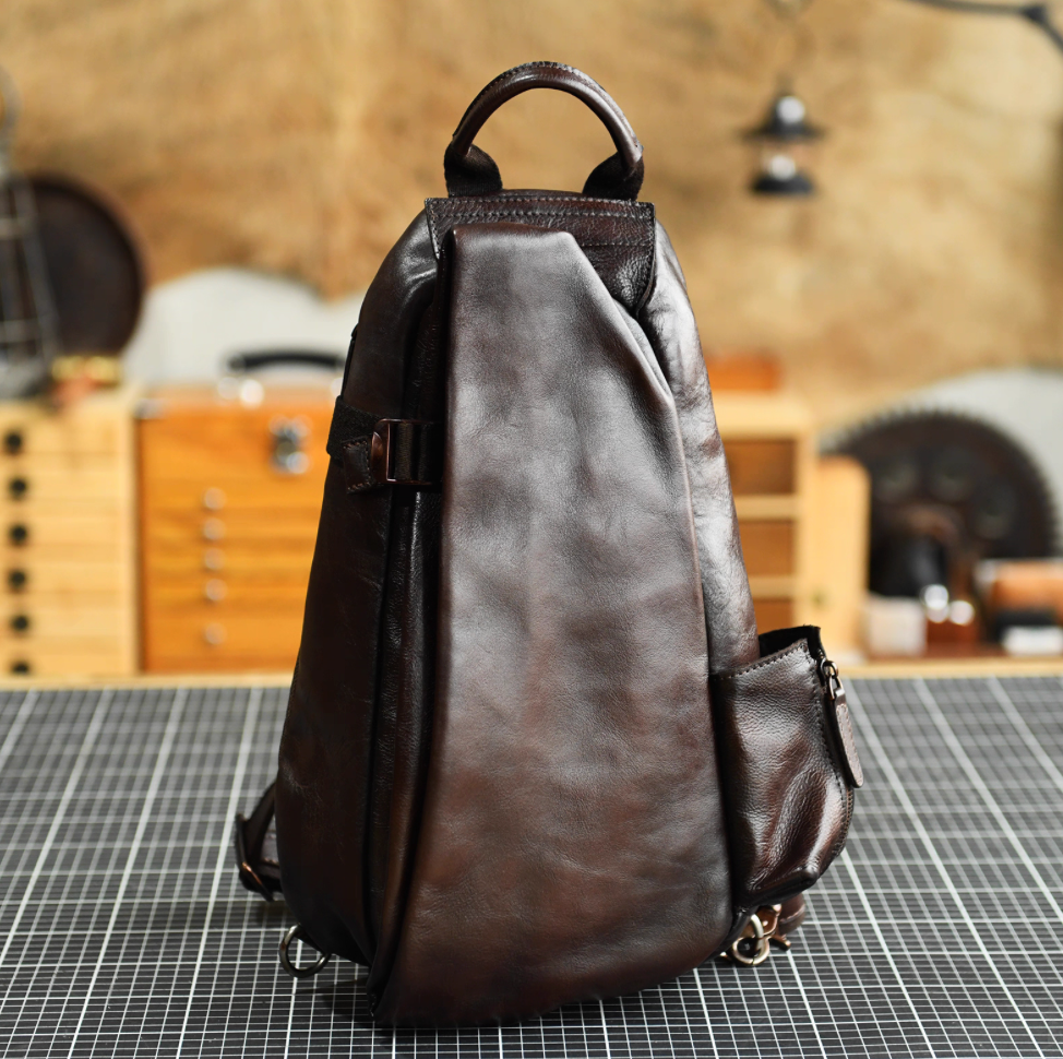Dark brown leather sling bag standing on worktable, single-handle top and side zip pocket.