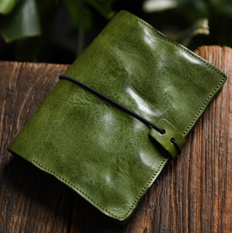Olive green full-grain leather refillable journal tied with black elastic on rustic wood.