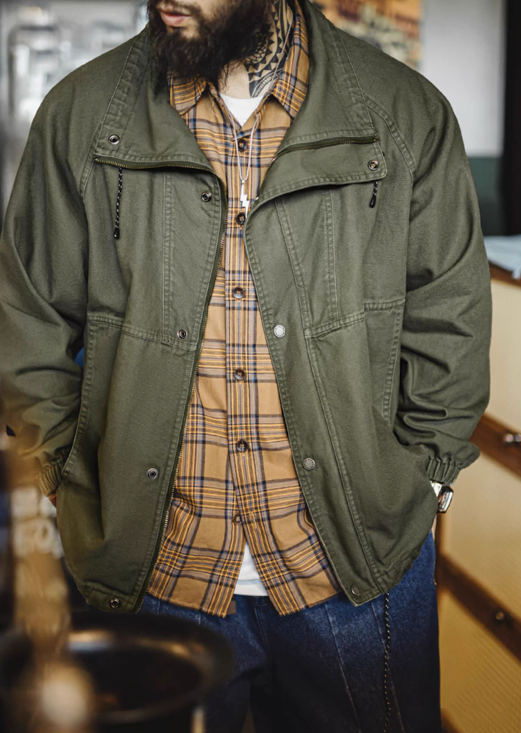 Model layering olive green M-65 jacket over plaid shirt for casual streetwear look