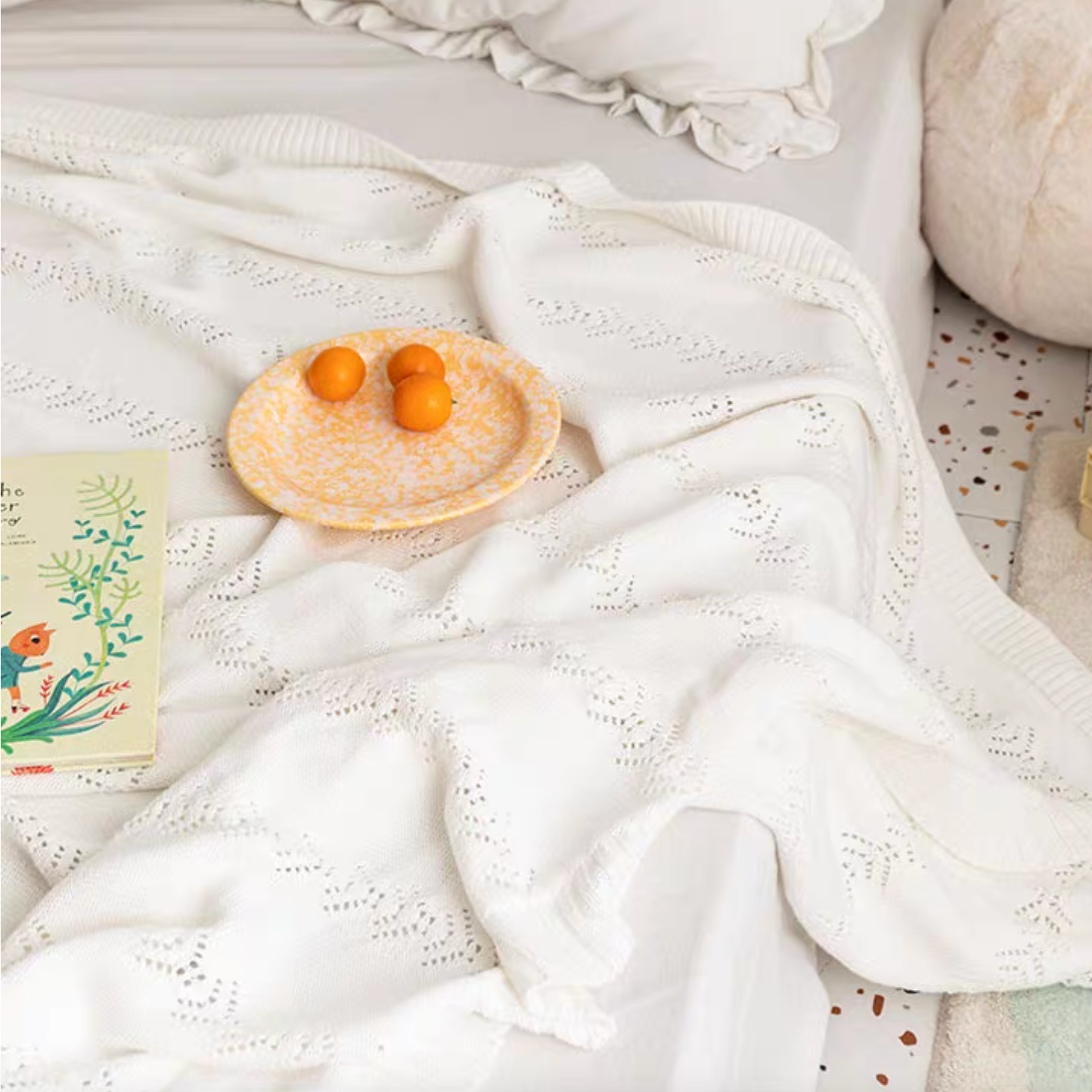 White Oversize Knit Throw Blanket draped across bed with plate of oranges and book highlighting breathable cotton knit