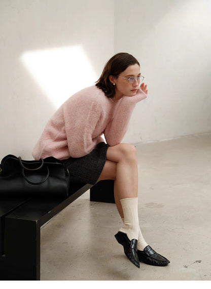 Pink alpaca sweater worn seated sideways under natural light, showing soft drape, long sleeves and casual styling with skirt and loafers