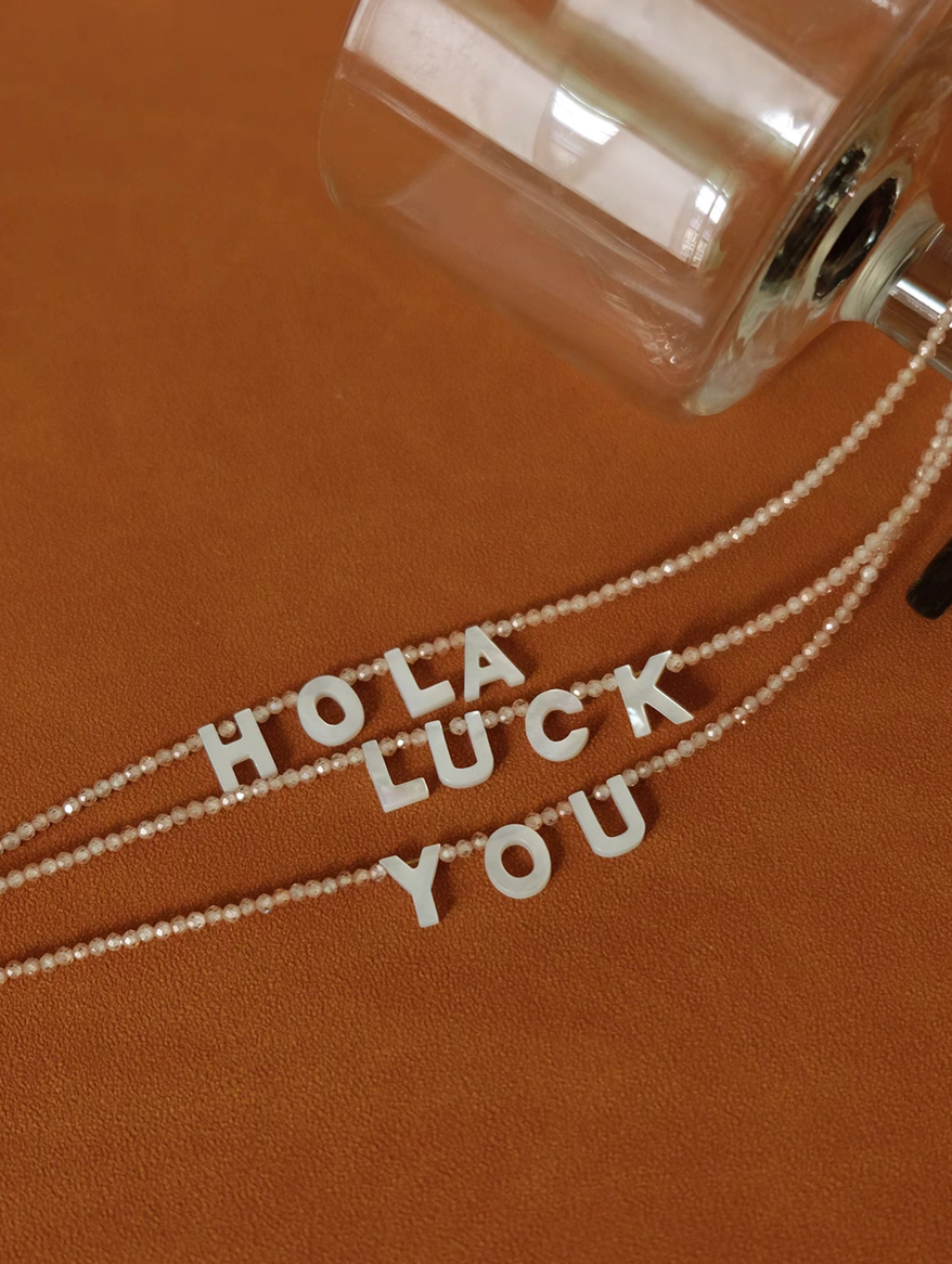 Personalized shell initial necklaces spelling HOLA LUCK YOU displayed on surface with glass detail and bead chains