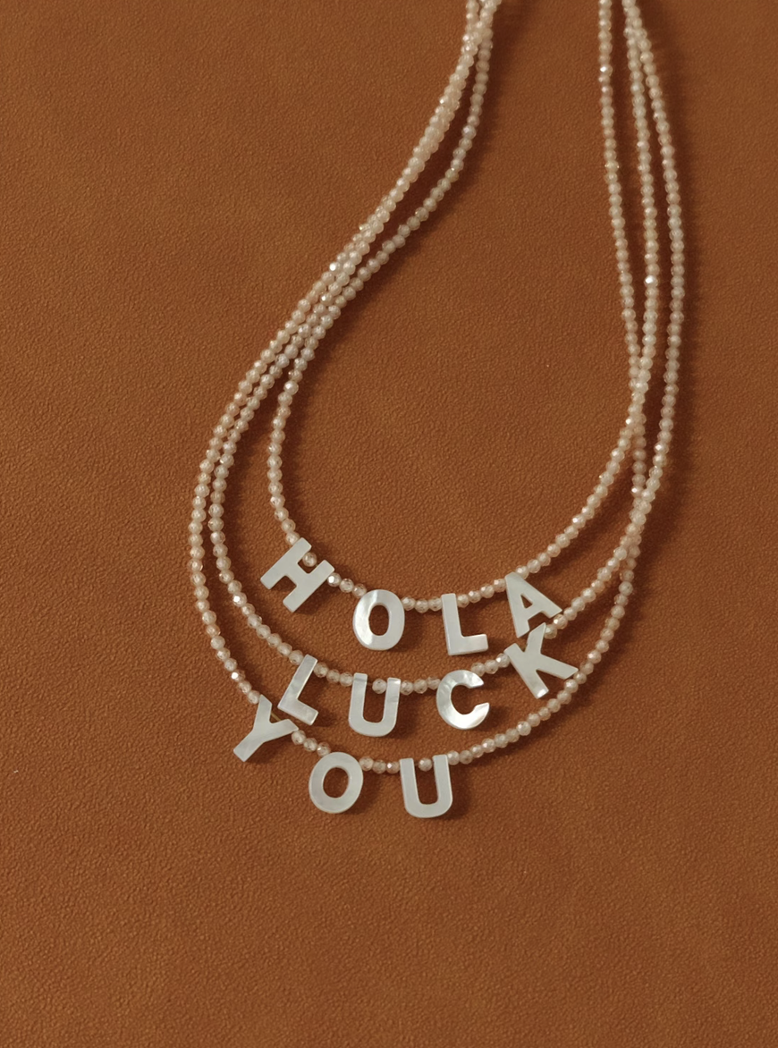 Layered personalized shell initial necklaces spelling HOLA LUCK YOU on beaded chains in natural color