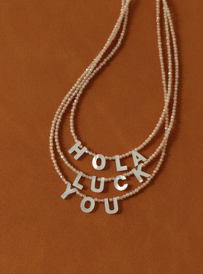 Layered personalized shell initial necklaces spelling HOLA LUCK YOU on beaded chains in natural color