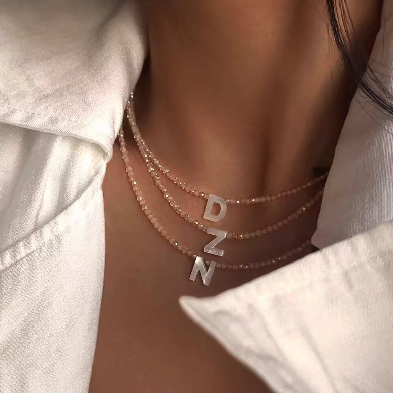 Layered personalized shell initial necklaces with letters D Z N worn on neck featuring natural bead chains