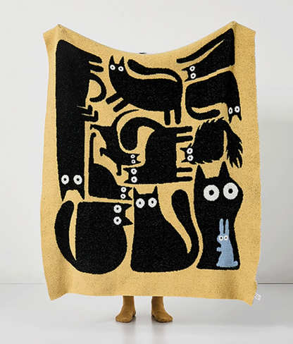 Yellow throw blanket held up fully displaying black abstract cats and a small blue figure with bold graphic layout