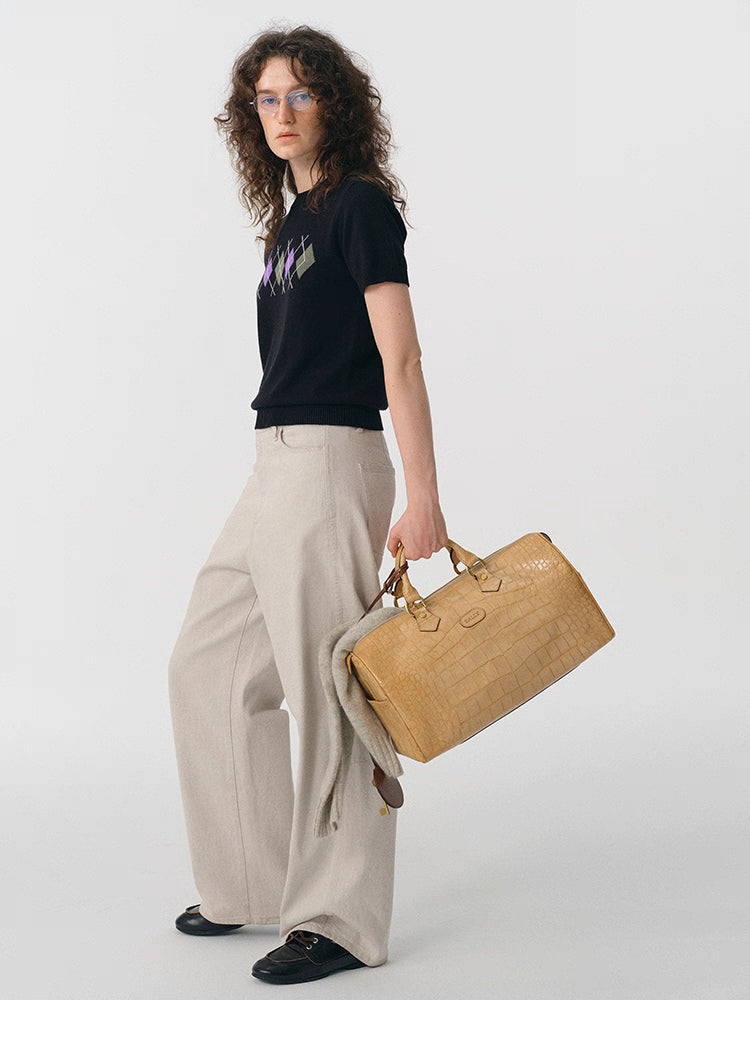 Model wearing black argyle knit tee carrying tan textured duffle bag.