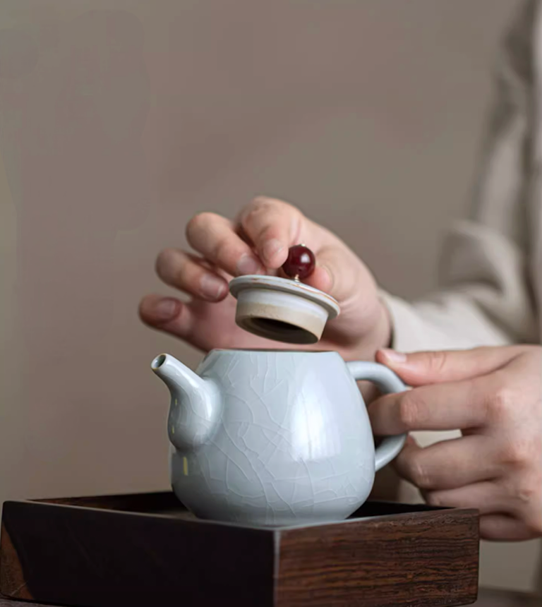 Ru Kiln Celadon Mini Teapot in light blue with lid being lifted showing opening detail and crackle glaze body on a wooden tray