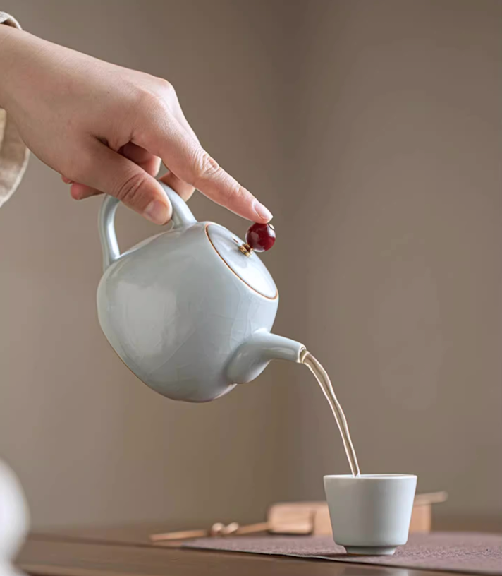 Ru Kiln Celadon Mini Teapot in light blue pouring tea into a small teacup showing steady stream and curved spout