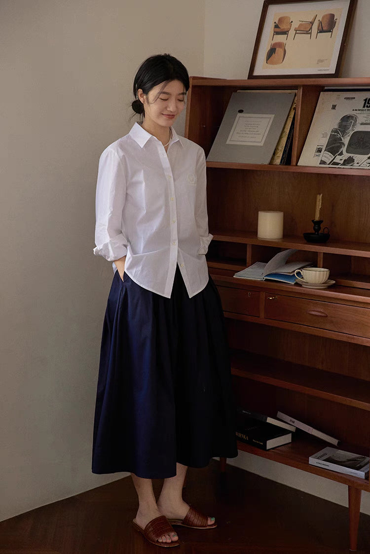 Model wearing The Sculptural Pleat A-Line Skirt in navy blue paired with white shirt showing elegant high waist and structured pleated silhouette