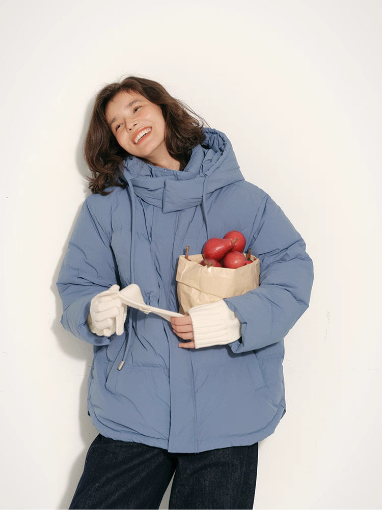 Woman wearing dusty blue detachable-hood puffer jacket smiling and holding apples in both hands.