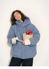 Woman wearing dusty blue detachable-hood puffer jacket smiling and holding apples in both hands.