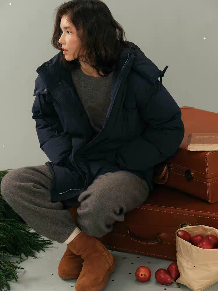 Model sitting beside brown luggage wearing navy blue puffer jacket in a relaxed side pose.