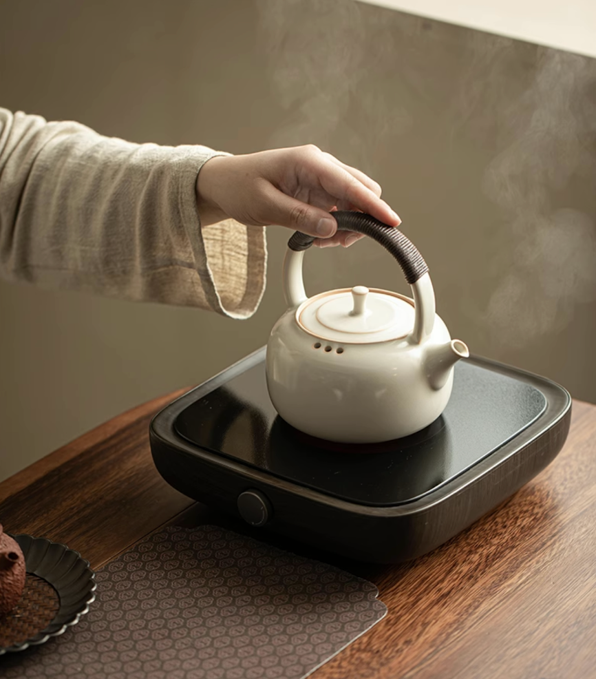 Soda Glaze Ceramic Teapot in off-white placed on an electric ceramic stove with hand holding the handle and steam rising
