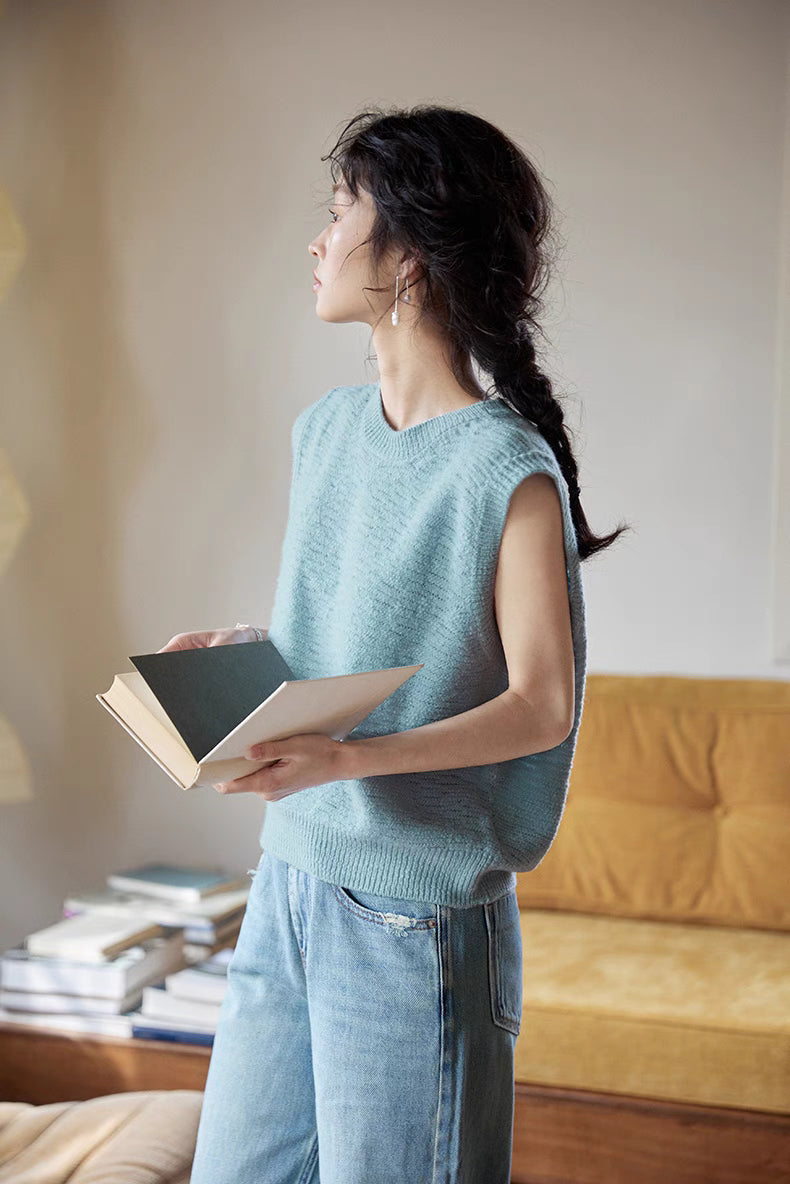 Model wearing Lake Blue merino sleeveless vest in side view holding a book, showcasing textured knit, soft drape, and relaxed fit silhouette