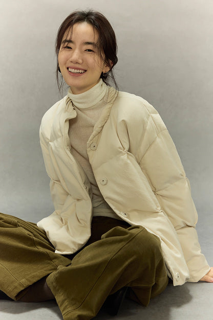 Model sitting cross-legged in cream beige SoftCloud down jacket, showing relaxed open front and layered knit underneath.