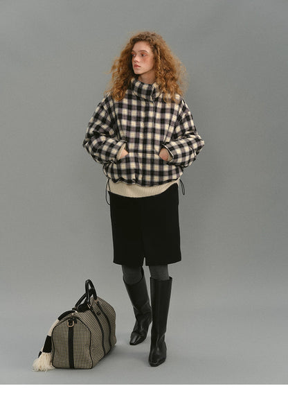 Front view of the SoftGrid wool check puffer zipped to a high stand collar, paired with a cream knit, black skirt, tall boots, and a check weekender bag on the floor.