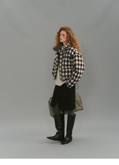 Full-body long shot of the SoftGrid wool check puffer styled with a black skirt, gray tights, and tall black boots, travel bag placed behind.