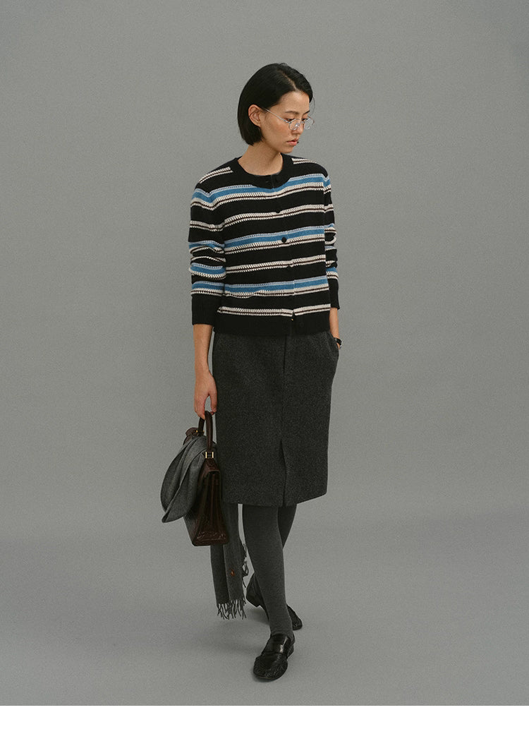 Model wearing the black-blue striped cardigan with a charcoal wool skirt, grey tights, and loafers, holding a bag and scarf.