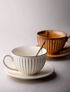 Striped ceramic cappuccino art cup and saucer in white with spoon shown in lifestyle setting alongside yellow variant in background