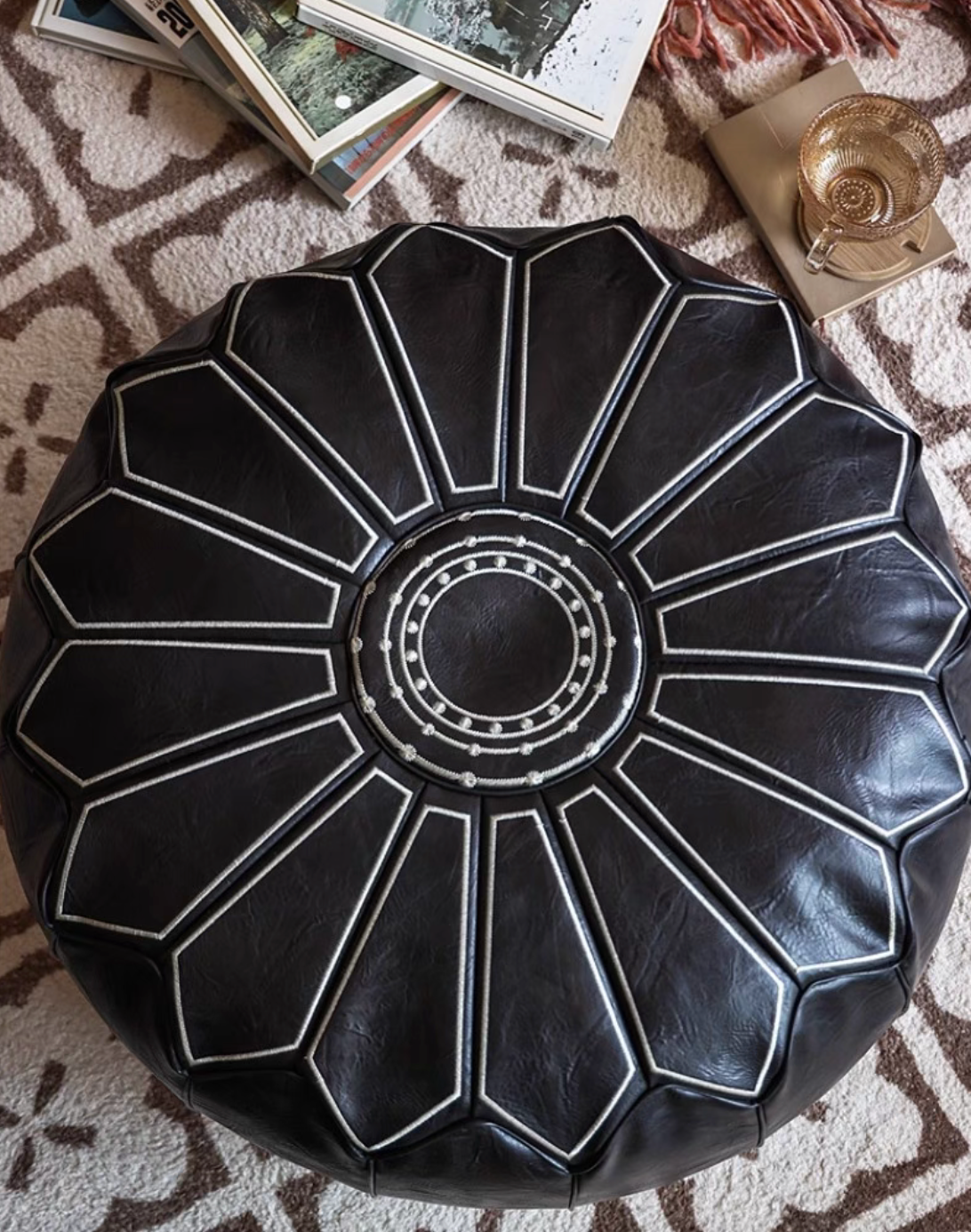 Top view of black faux leather pouf ottoman with books and cup on patterned rug