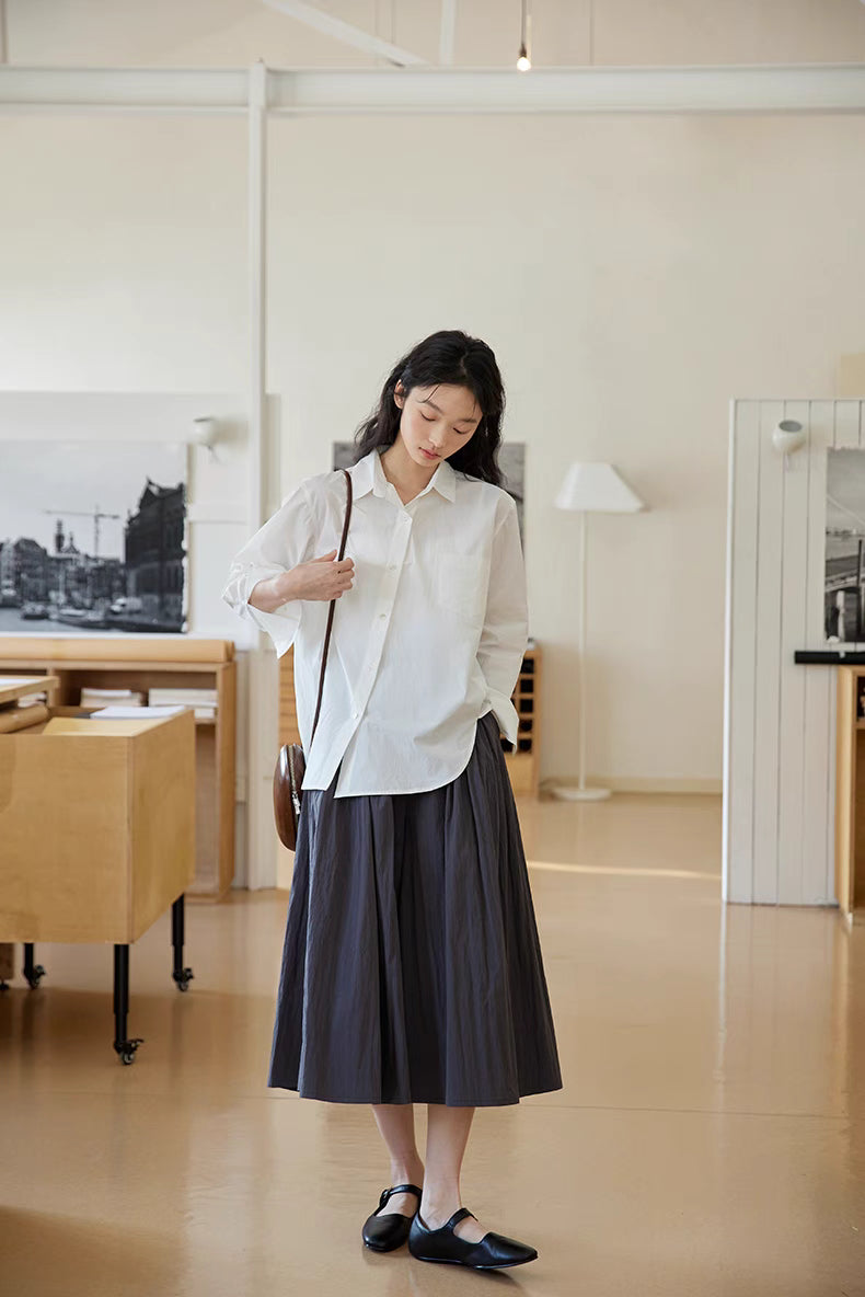 Model wearing The Poised Pleated Midi Skirt in charcoal gray paired with a white shirt, showing full-body front view with A-line silhouette and soft pleats