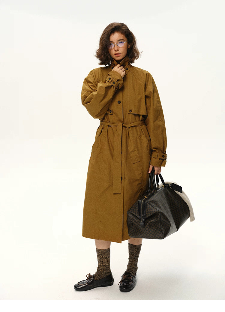 Woman wearing a maple brown cotton long trench coat standing front-facing with bag.
