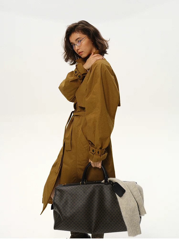 Side view of woman in maple brown trench coat holding a black travel bag.