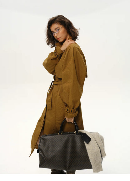 Side view of woman in maple brown trench coat holding a black travel bag.