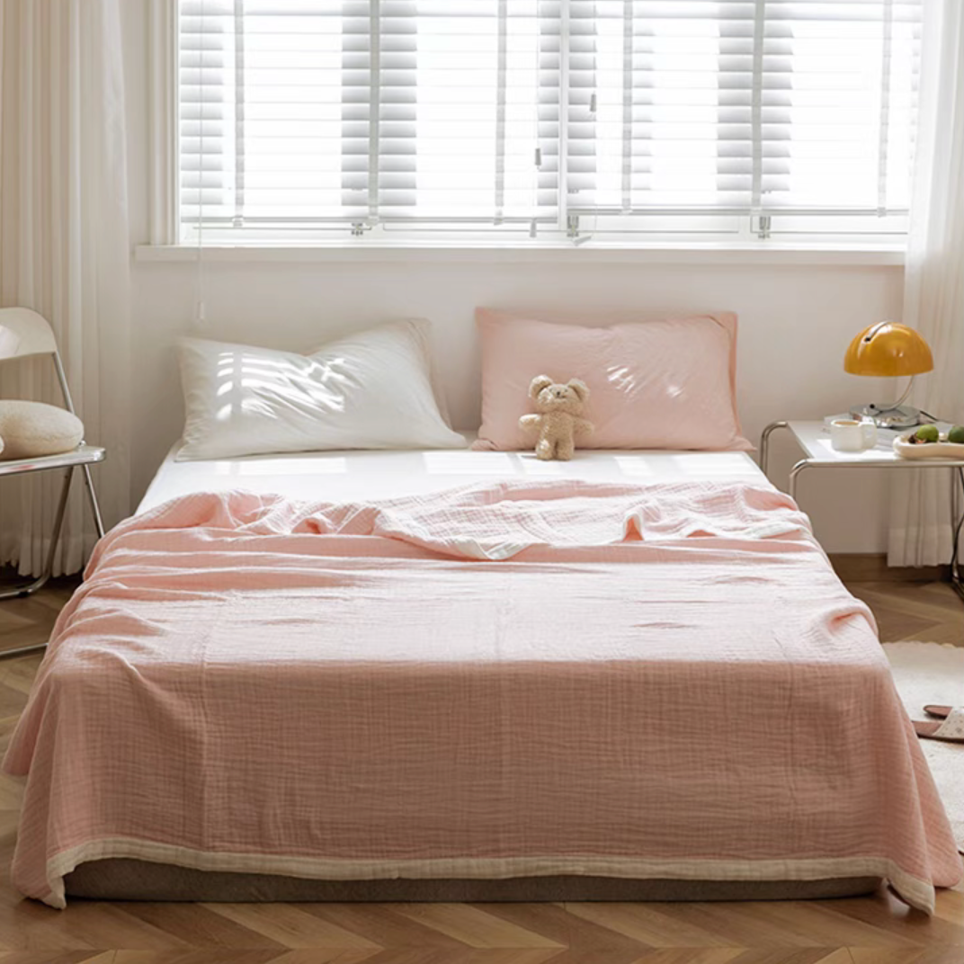 Light pink ripple gauze cotton quilt draped on bed with teddy bear decoration in sunlit bedroom.