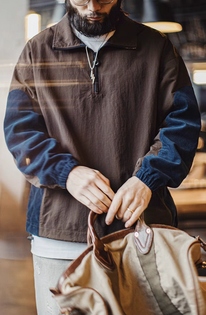 Male model wearing the URBANCORE dark coffee brown half-zip patch jacket in a café setting, paired with neutral trousers and travel bag.