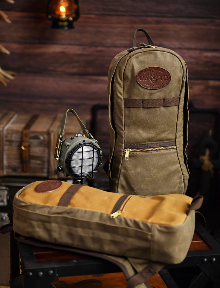 Two U.S. Mail waxed canvas backpacks displayed on a wooden table beside vintage lantern and props.