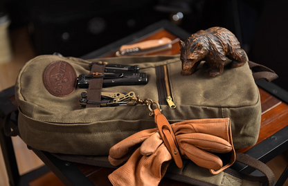 Army green waxed canvas backpack laid flat with gloves and multitools beside wooden bear ornament.