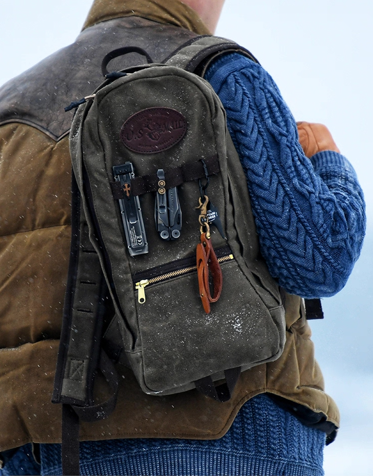 Man wearing U.S. Mail waxed canvas backpack outdoors in snowy weather with brown vest and blue sweater.