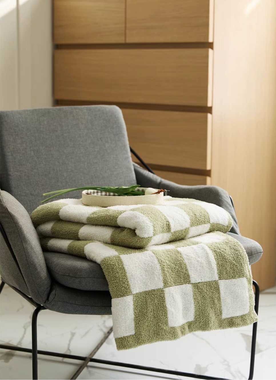 Matcha green checkered blanket folded on gray lounge chair with tray and greenery placed on top