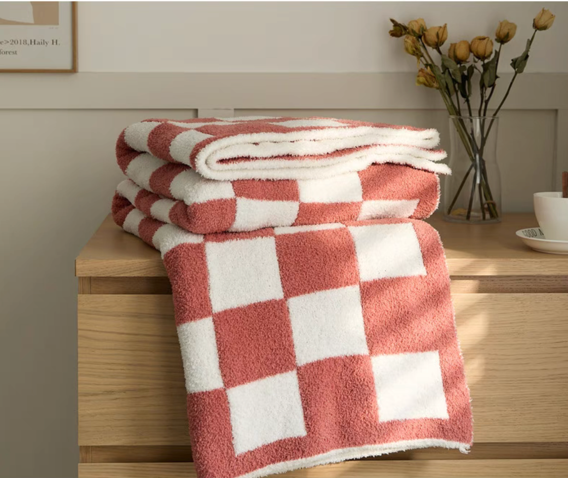 Folded watermelon red checkered blanket stacked on wooden table with vase of dried flowers beside