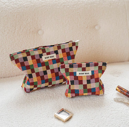 Two sizes of Vibrant Checkered Travel Makeup Bag in multi-colored checkered pattern placed on soft fabric surface with beauty accessories nearby