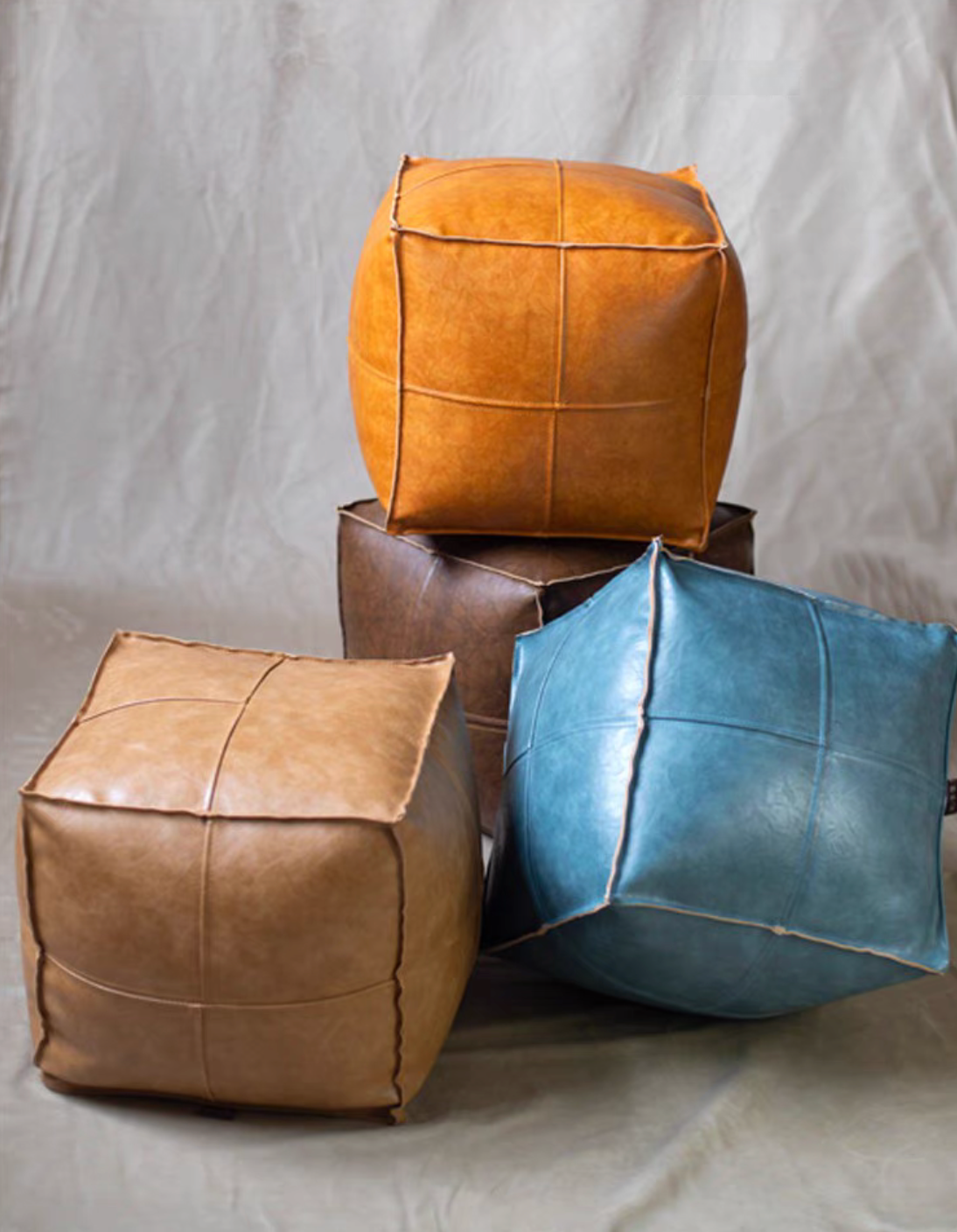 Faux leather square floor poufs in orange, dark brown, tan, and blue stacked together showing clean stitched panel design